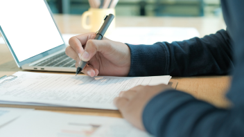 Person signing paperwork beside an open laptop